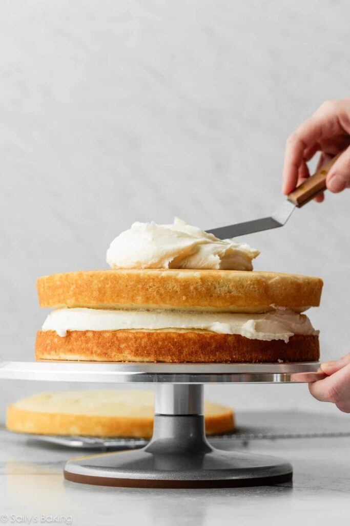 Layered vanilla cake being frosted on a stand with a spatula for smooth finishing.