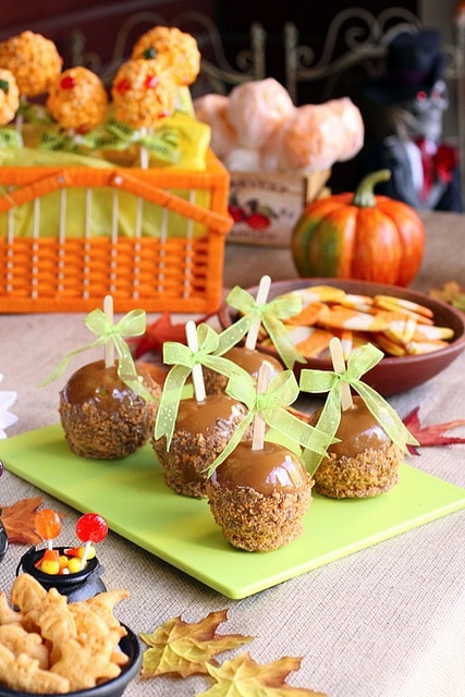 Caramel apples with green ribbons on platter, surrounded by fall treats and pumpkins. Perfect for autumn gatherings.