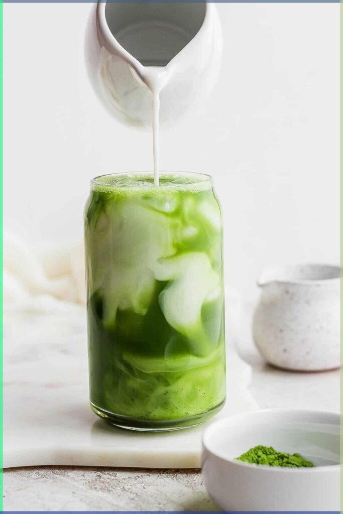 Creamy milk pouring into a glass of iced matcha latte, next to a bowl of matcha powder.