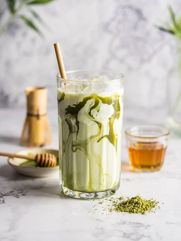Iced matcha latte in tall glass with bamboo straw, surrounded by matcha, honey, and bamboo whisk on a marble surface.