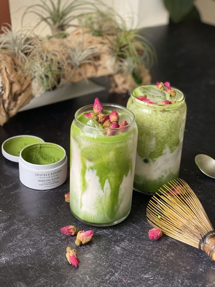 Iced matcha lattes with rose buds on a table, accompanied by a bamboo whisk and matcha powder tin.