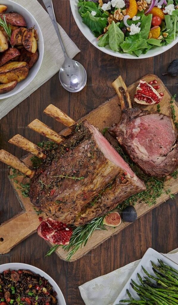 Roasted bone-in prime rib on a wooden board with pomegranate, surrounded by salad, potatoes, and asparagus.