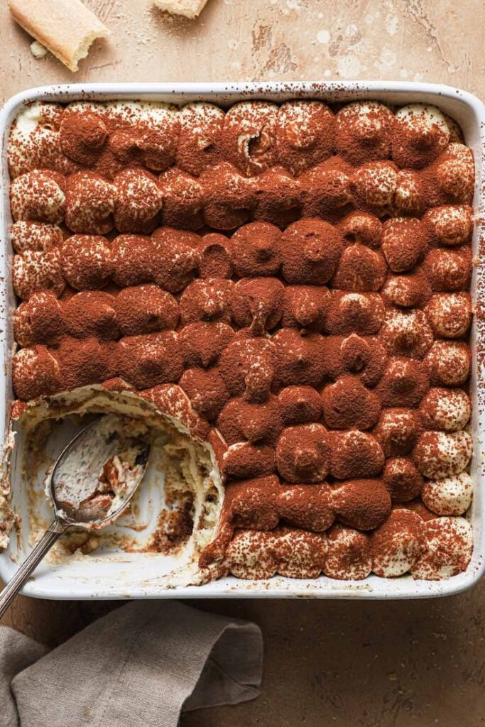 Tiramisu dessert in a baking dish with cocoa powder, one scoop removed, and a spoon beside it on a beige surface.