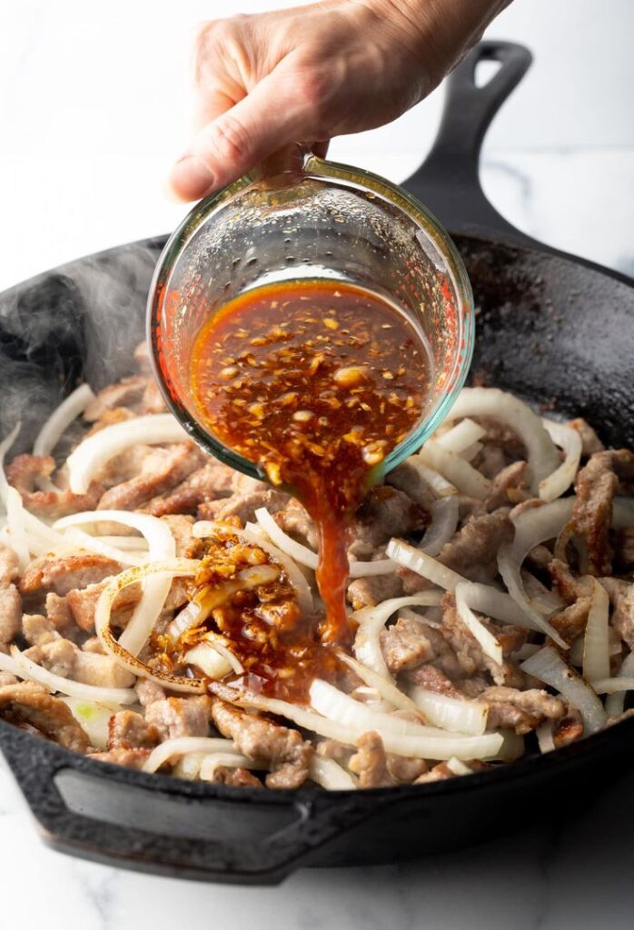 Pouring savory sauce over sizzling onions and meat in a cast-iron skillet for a flavorful stir-fry dish.