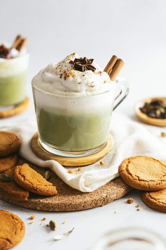 Creamy matcha latte topped with frothy whipped cream, cinnamon, and star anise, served with cookies on a cozy setup.