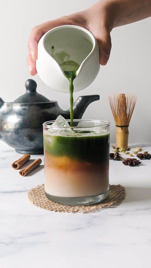 Pouring matcha into a layered iced latte with teapot, whisk, and spices in the background on a marble surface.