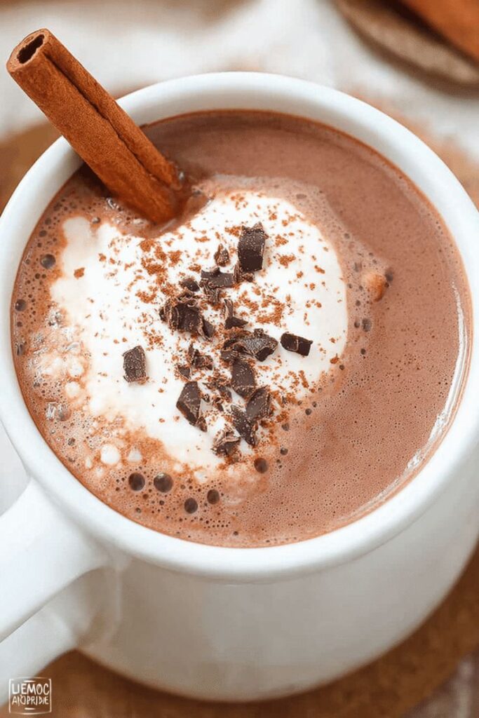Creamy hot chocolate topped with whipped cream, chocolate shavings, and cinnamon stick in a white mug.