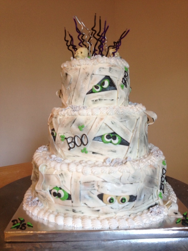 Three-tier Halloween-themed cake with mummy eyes, Boo text, and curly ribbon decorations.