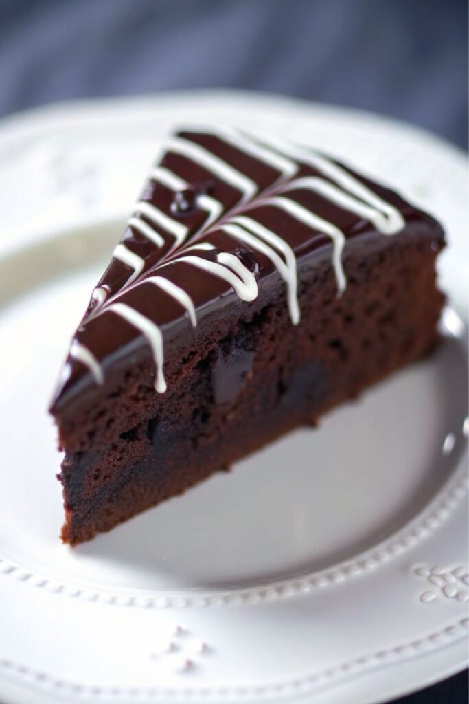 Rich chocolate cake slice with dark glaze and white icing drizzle on a white plate.
