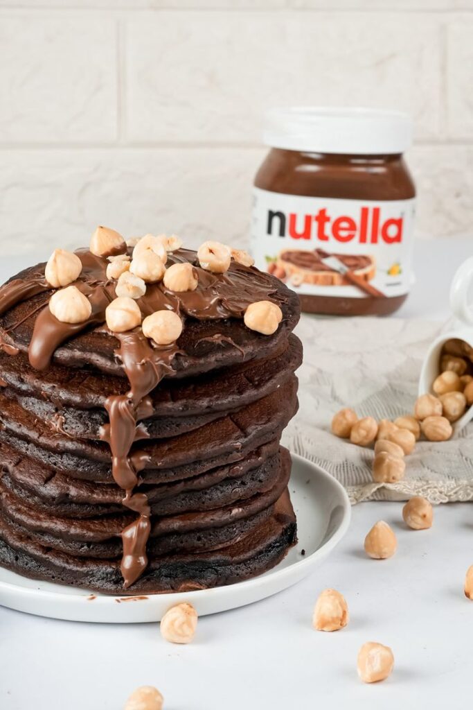 Stack of chocolate pancakes drizzled with Nutella and topped with hazelnuts, with a Nutella jar in the background.