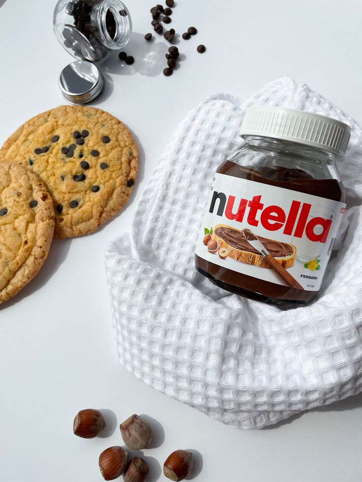 Jar of Nutella with chocolate cookies and hazelnuts on a white cloth, surrounded by chocolate chips.