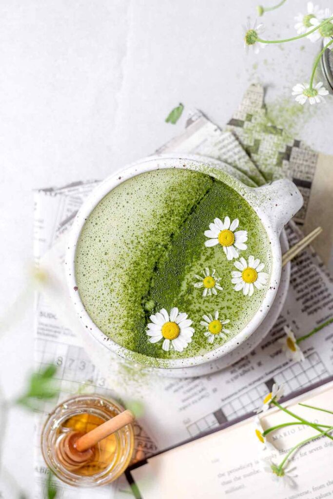 Ceramic mug of vibrant matcha latte garnished with chamomile flowers, surrounded by honey and papers.
