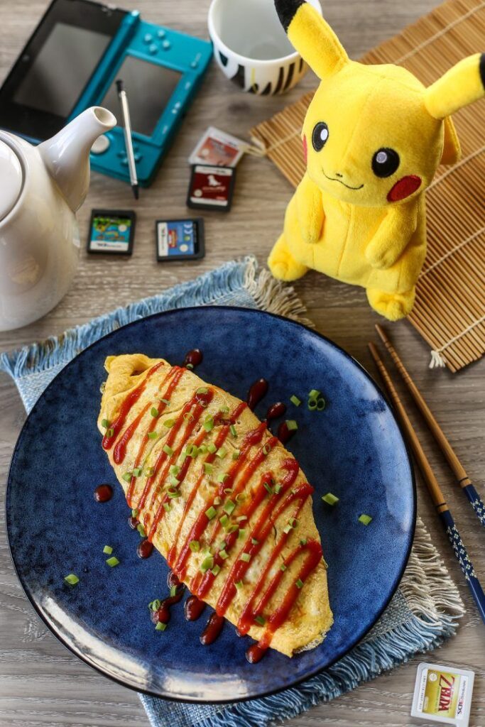 Omelette drizzled with sauce on blue plate, next to a gaming console and plush toy on a wooden table.