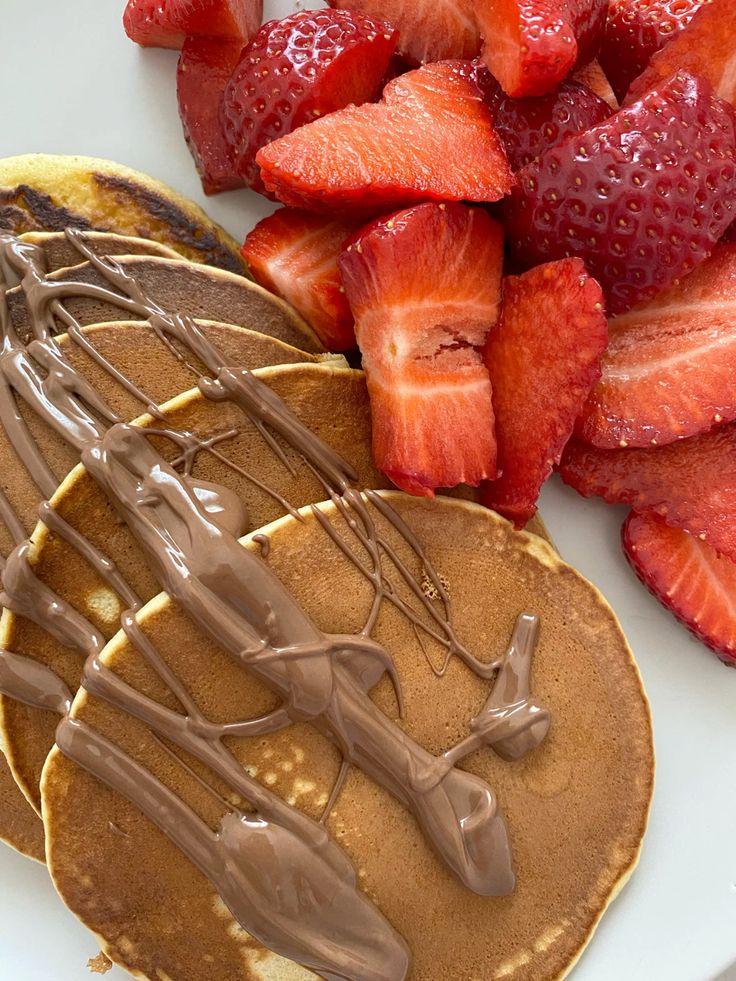 Pancakes topped with chocolate syrup served with fresh sliced strawberries on a white plate.