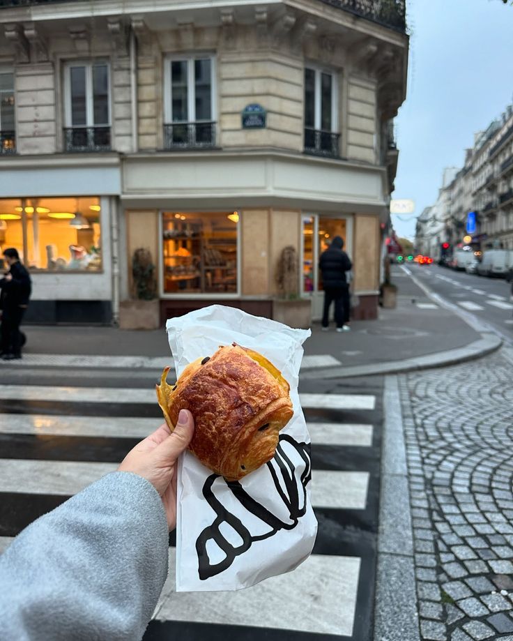 Hand holding a chocolate croissant in front of Parisian bakery on a street corner.