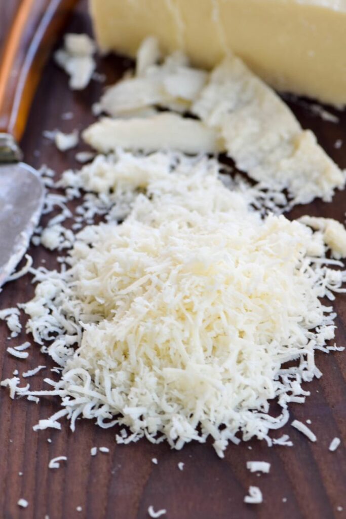 Grated parmesan cheese on a wooden board with a knife and cheese block in the background.