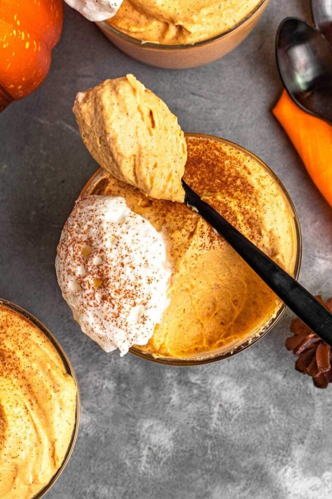 Creamy pumpkin mousse topped with whipped cream and cinnamon in a glass, served with a spoon on a gray table.