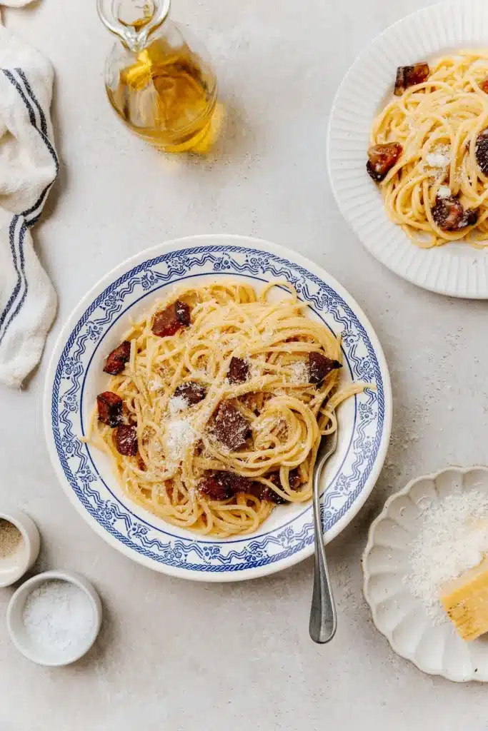Plate of spaghetti carbonara with grated cheese, crispy bacon, and olive oil on a light table setting.