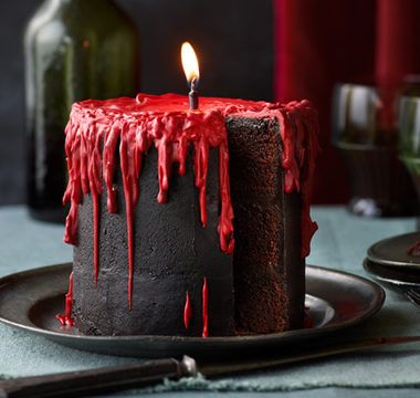 Dark cake with red drips, lit candle on top, displayed on a plate, creating a gothic, mysterious atmosphere.