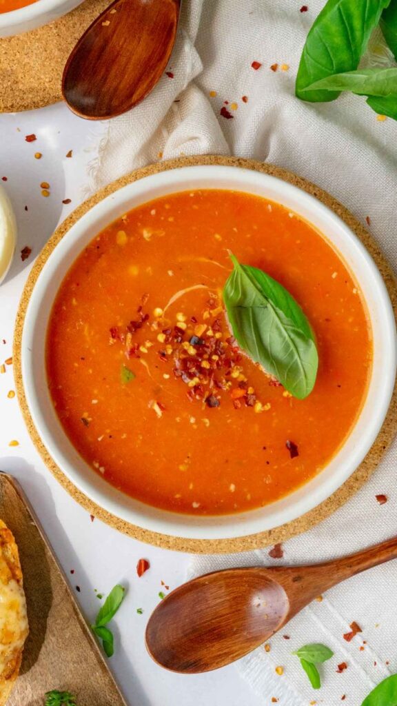 Tomato soup garnished with basil and chili flakes in a white bowl, with a wooden spoon beside it.