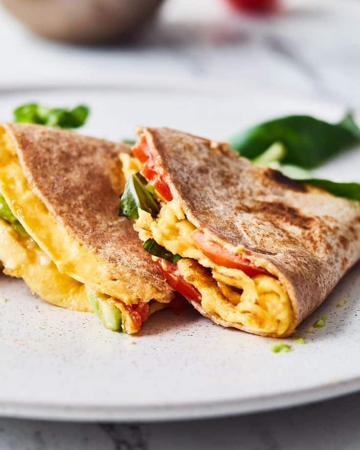 Delicious breakfast tortilla wrap filled with scrambled eggs, fresh vegetables, and cheese on a white plate.