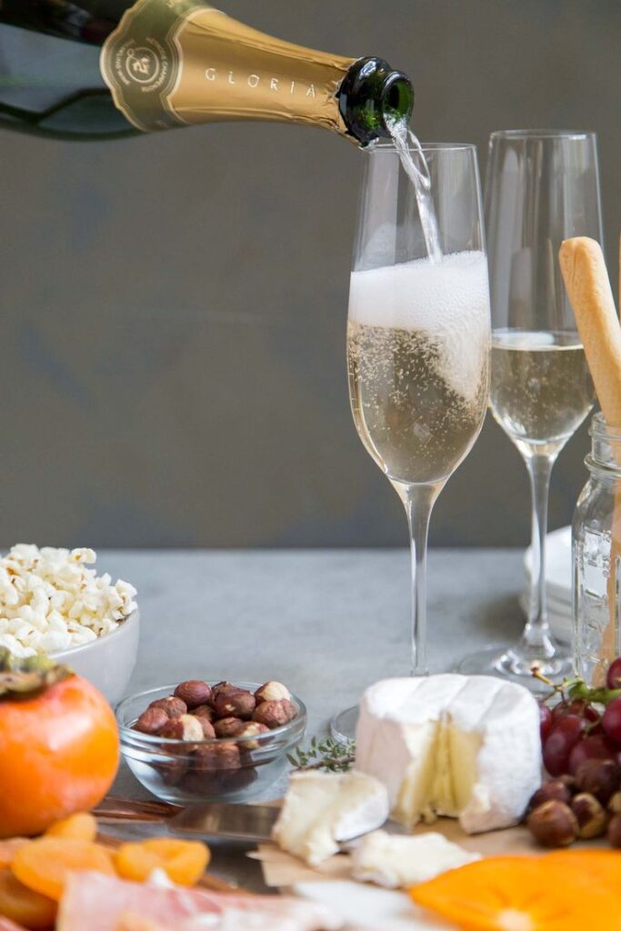 Sparkling Wine Food Pairing- The Little Epicurean - 10 Crowd-Pleasing Appetizers That Will Wow Your Party | Pouring champagne into a flute with a gourmet charcuterie spread of cheese, nuts, popcorn, and fruit.