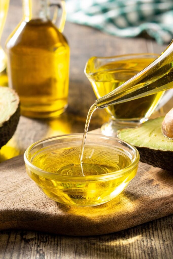 Pouring fresh avocado oil into a glass bowl on a rustic wooden table, with whole avocados in the background.