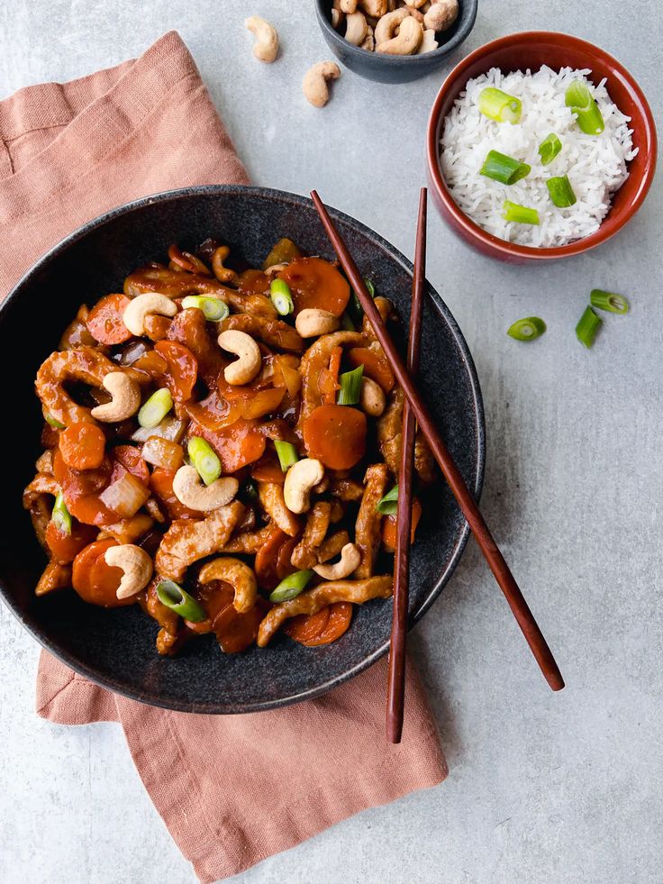 Stir-fried chicken with cashews, carrots, and scallions in black bowl, served with rice and chopsticks on side.