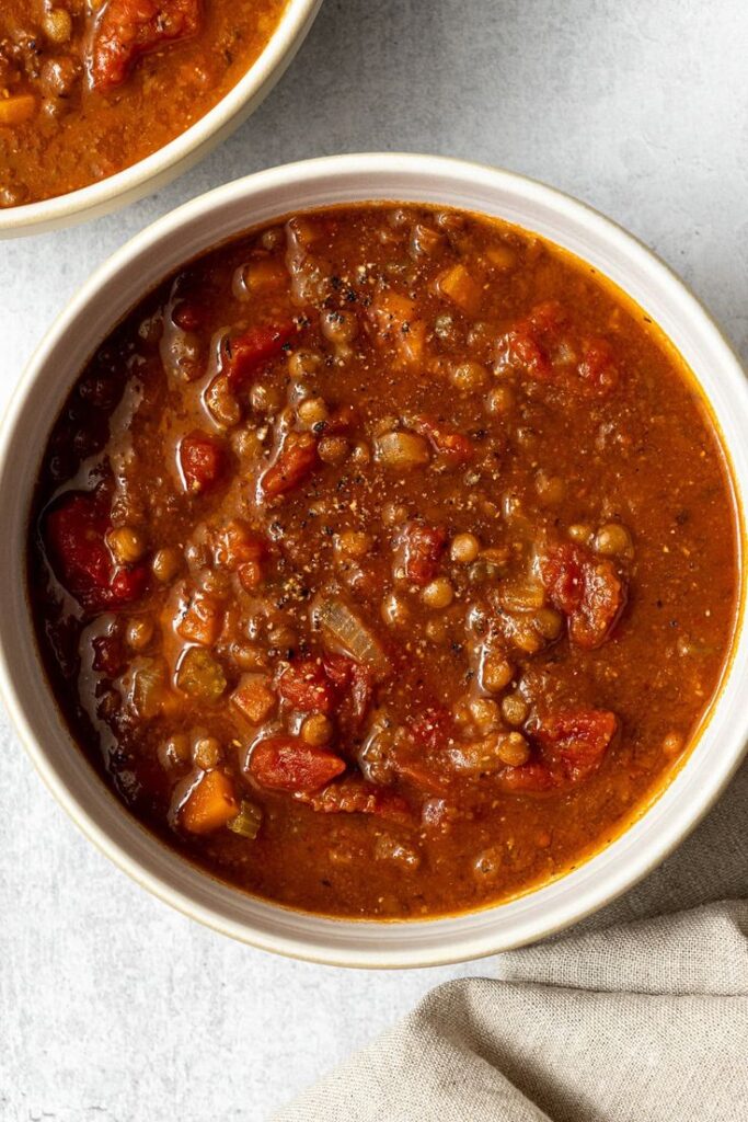 Hearty vegetarian lentil soup in a white bowl, filled with tomatoes and spices, on a textured background.