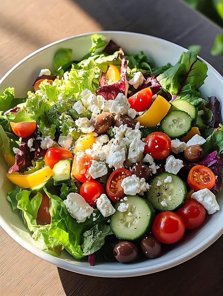 Fresh Greek salad with lettuce, cherry tomatoes, cucumber, olives, feta cheese, and peppers in a white bowl.