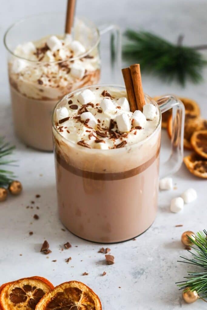 Two mugs of hot chocolate topped with whipped cream, marshmallows, cinnamon sticks, and chocolate shavings. Cozy winter treat.
