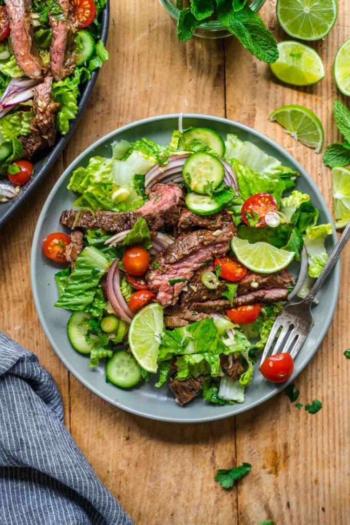Fresh steak salad with lime, cherry tomatoes, cucumbers, and greens on a plate, garnished with mint leaves.