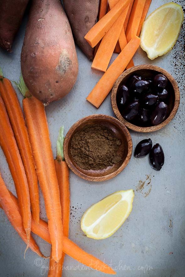 Carrots, sweet potatoes, black olives, cumin, and lemon slices on a table.