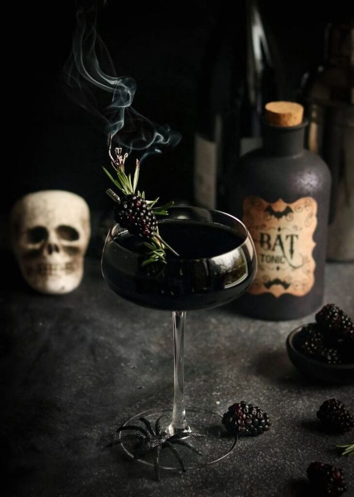 Spooky black cocktail with rosemary and blackberries, surrounded by skull and decorative spider for Halloween vibe.