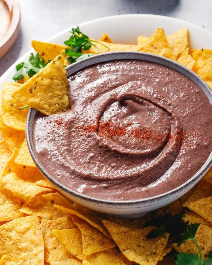 Bowl of creamy black bean dip with tortilla chips and garnish, perfect for snacking or parties.