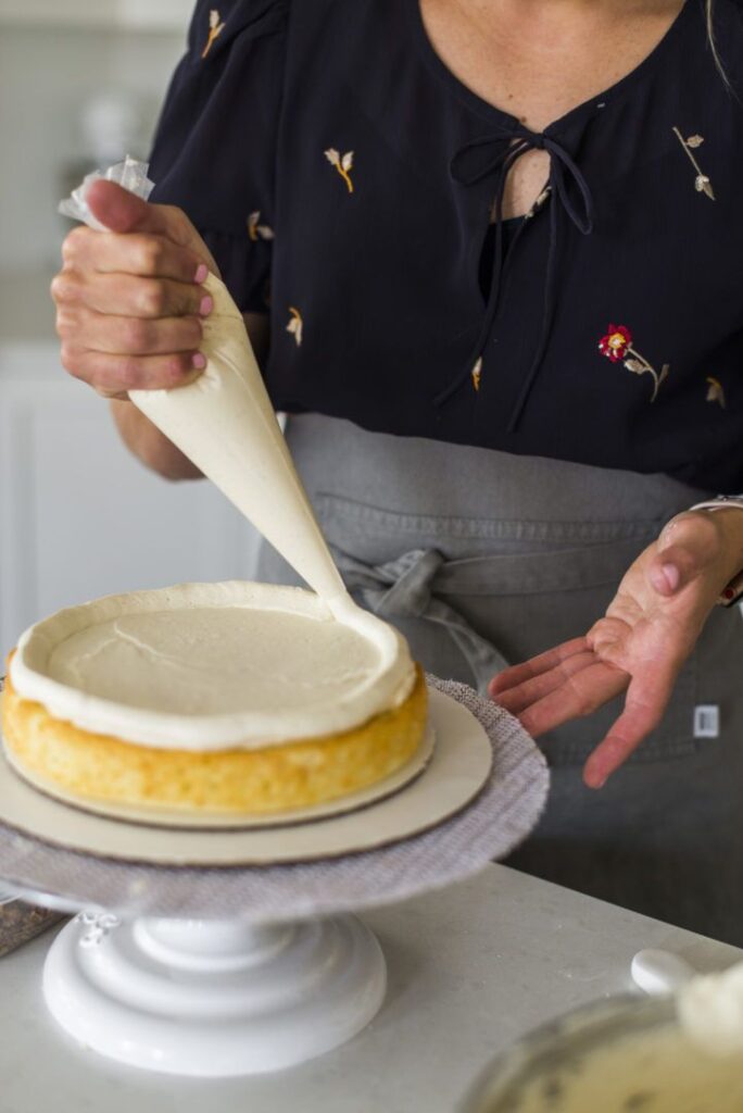 Person decorating cake with piping bag on stand in kitchen.