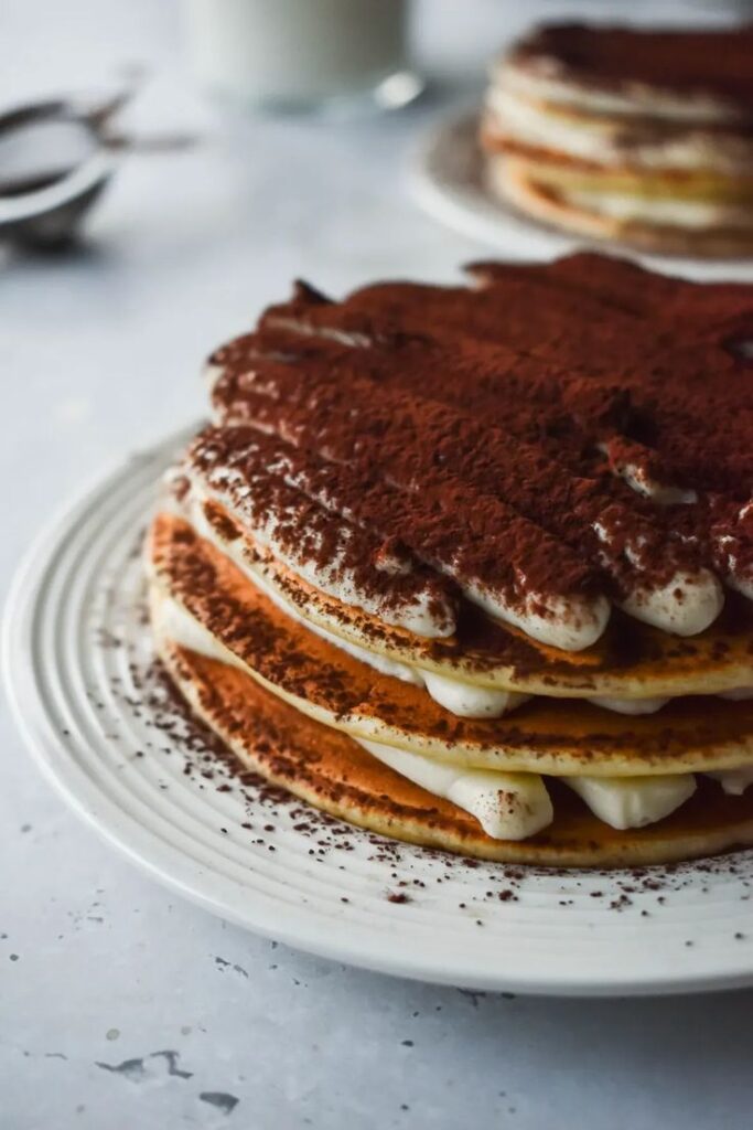 Tiramisu pancake stack topped with cocoa powder on a white plate, perfect for a delicious breakfast treat.