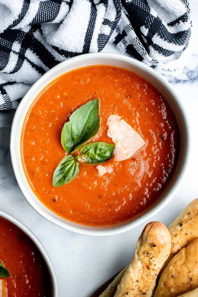 Bowl of creamy tomato soup with fresh basil and parmesan, served with breadsticks, on a marble countertop.