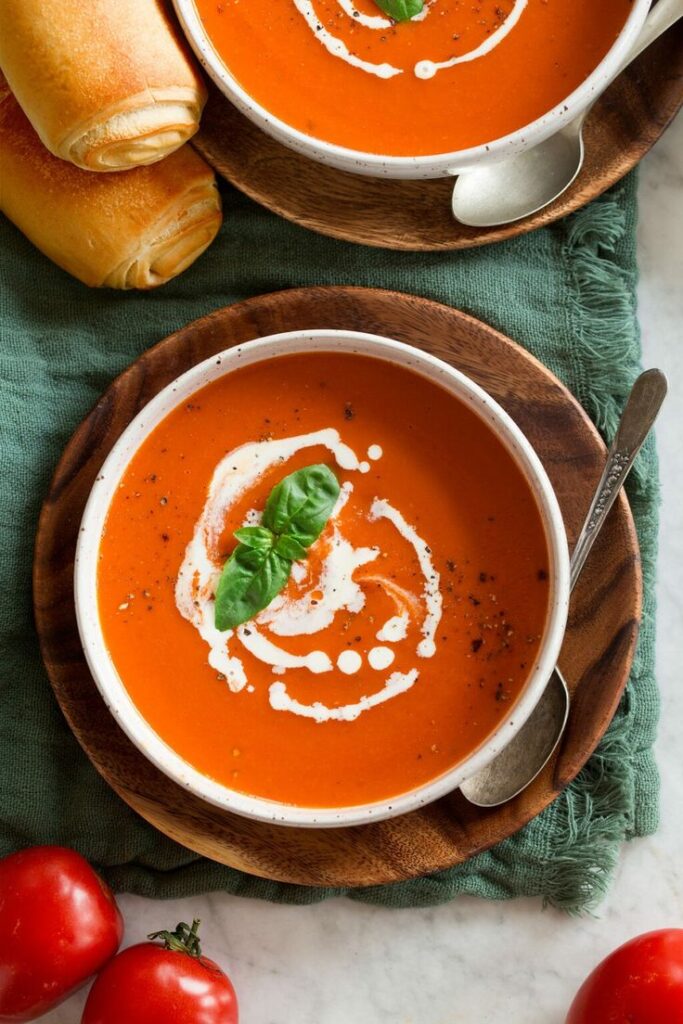 Creamy tomato soup with basil garnished and bread rolls on a wooden plate, perfect for a cozy meal.