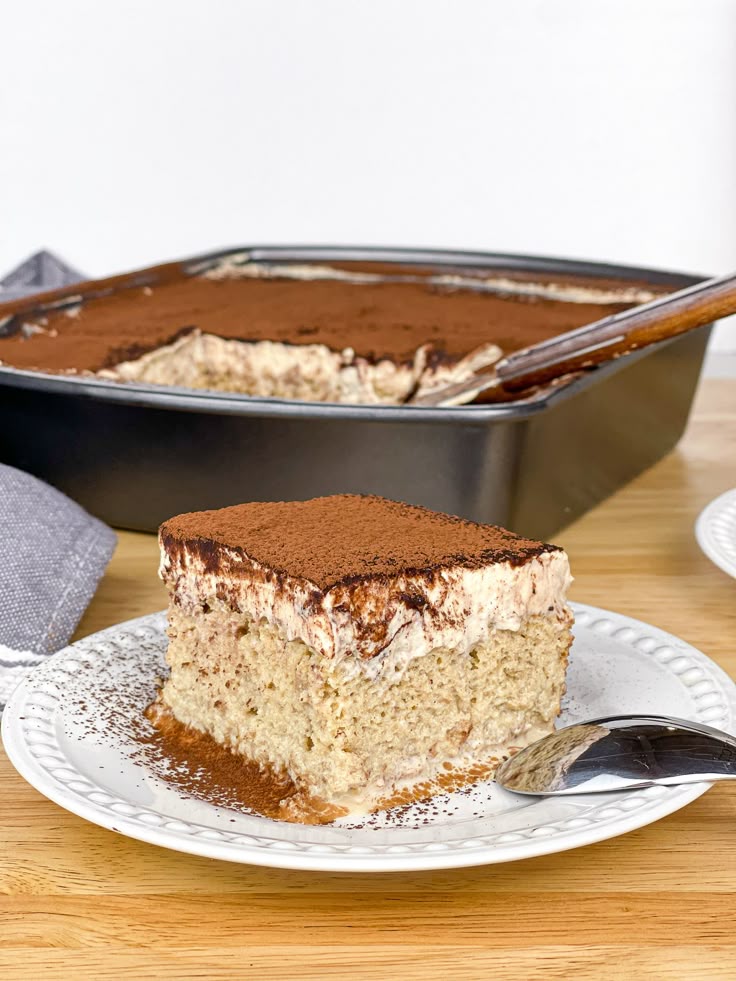 Slice of creamy tiramisu cake with cocoa topping on a white plate, placed beside a baking dish on a wooden table.
