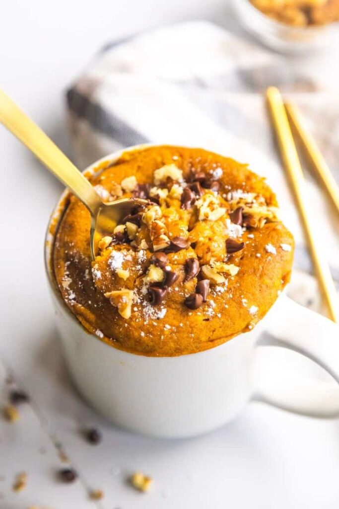 Pumpkin mug cake topped with chocolate chips and nuts in a white mug with a golden spoon. Perfect fall dessert.