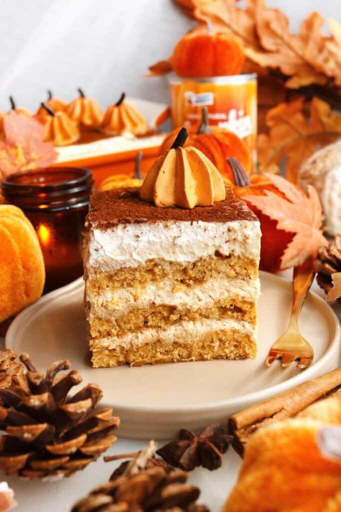 Slice of pumpkin tiramisu on a plate, garnished with whipped cream and surrounded by fall decor.