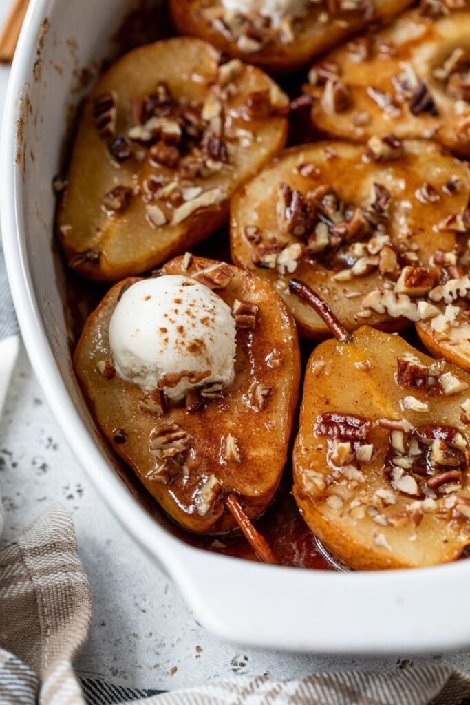Baked pears with pecans topped with ice cream in a white dish, featuring cinnamon spice and caramel drizzle.