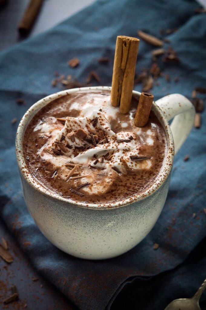 Creamy hot chocolate topped with whipped cream, chocolate shavings, and cinnamon sticks in a ceramic mug.
