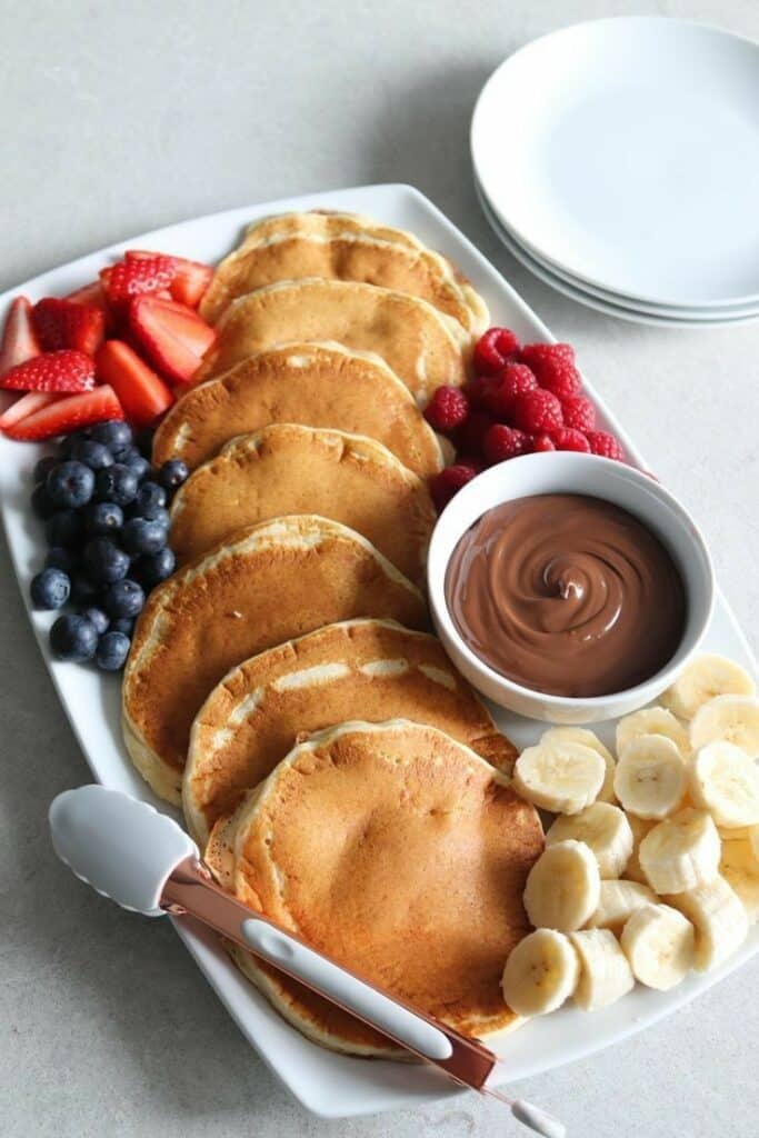 Pancakes with strawberries, blueberries, raspberries, bananas, and chocolate sauce on a platter for a delicious breakfast.