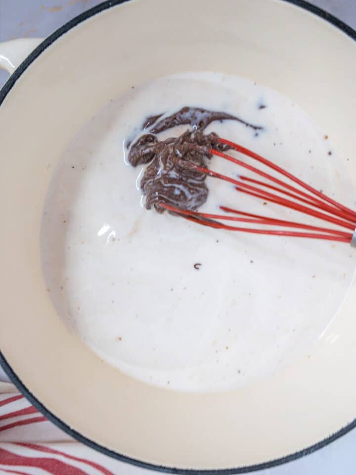 Mixing chocolate into creamy sauce in a pot with red whisk, cooking preparation.
