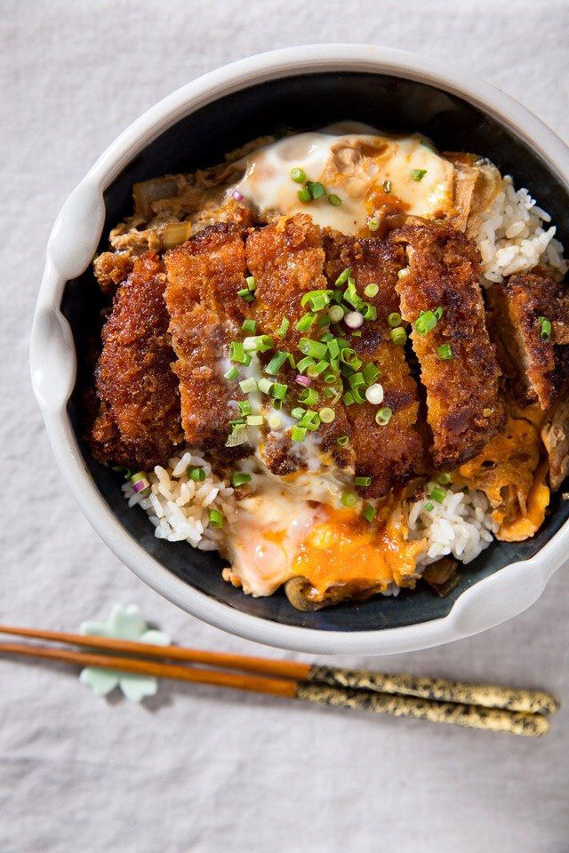 Bowl of crispy katsu on rice topped with egg and green onions, served with chopsticks.
