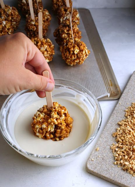 Dipping caramel popcorn ball into white chocolate with chopped nuts on a counter, perfect treat preparation.