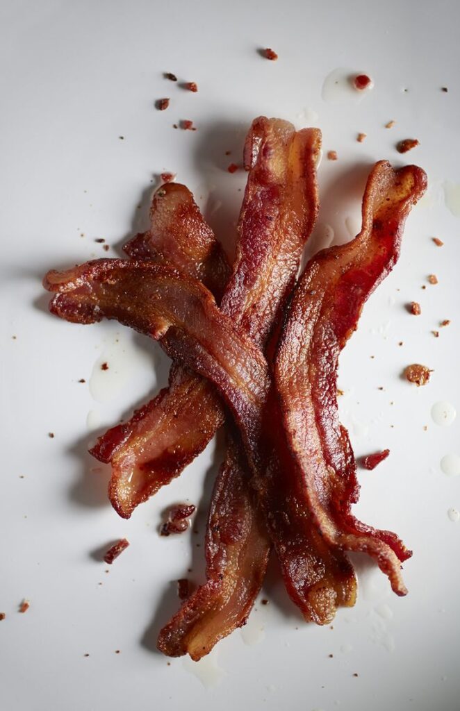 Crispy, golden-brown bacon strips with scattered crumbs on a white background, showcasing rich texture and savory appeal.