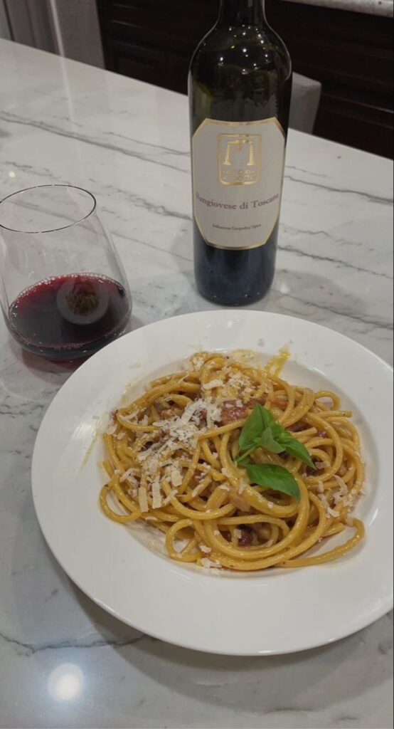 Plate of spaghetti with basil and cheese, paired with red wine bottle and glass on marble table.